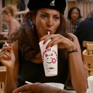 May include: A woman wearing a black beret and a black top is drinking from a white cup with the text "Teriyaki Dono" and a cartoon cat illustration. She is holding a lit cigarette with red nail polish.