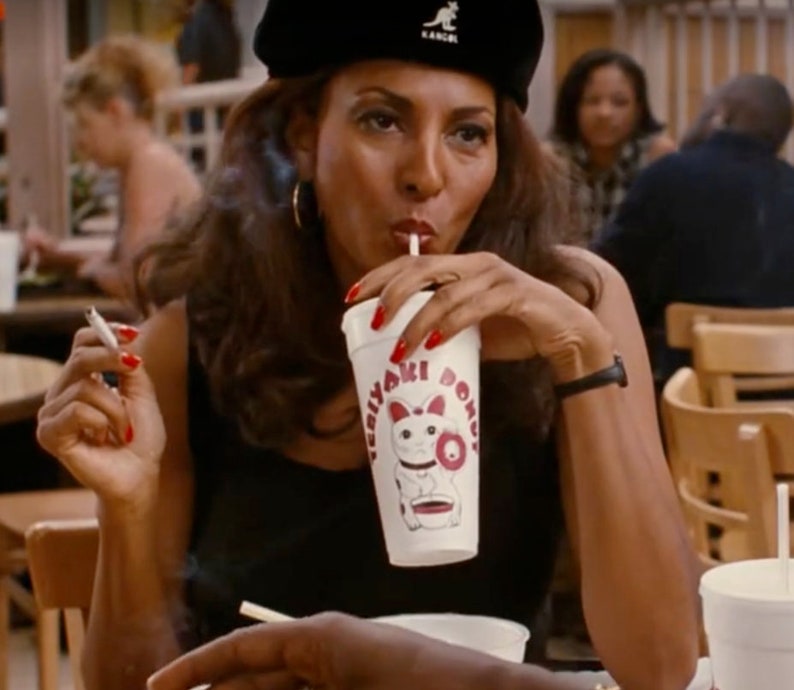 May include: A woman wearing a black beret and a black top is drinking from a white cup with the text "Teriyaki Dono" and a cartoon cat illustration. She is holding a lit cigarette with red nail polish.