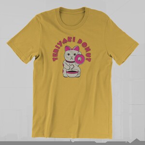 May include: Mustard yellow t-shirt featuring a cartoon cat holding a pink donut and a cup. The text "TERIYAKI DONUT" arches above the cat in pink letters. The cat has gray fur with pink accents and a serious expression.