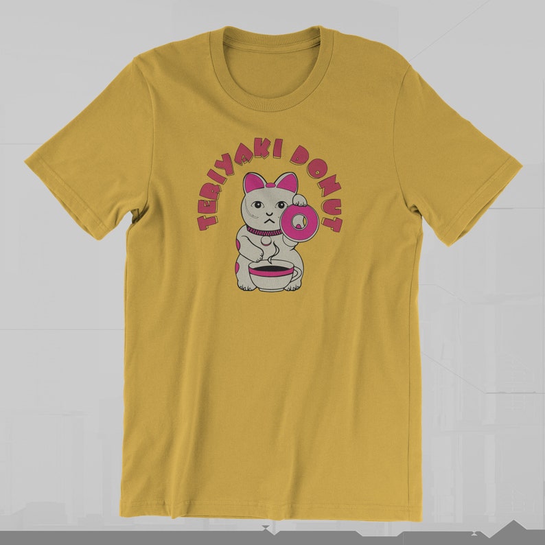 May include: Mustard yellow t-shirt featuring a cartoon cat holding a pink donut and a cup. The text "TERIYAKI DONUT" arches above the cat in pink letters. The cat has gray fur with pink accents and a serious expression.