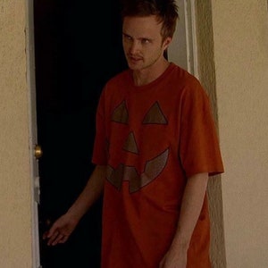 Może przedstawiać: Mężczyzna w pomarańczowym t-shircie z brązowym wzorem twarzy dyni. Stoi w drzwiach i trzyma klamkę.