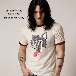 May include: A vintage white ringer t-shirt with dark-red trim on the collar and sleeves. The center features a distressed graphic of a black cat head with a wide grin, large eyes, and a long, bright red tongue hanging out of its mouth.