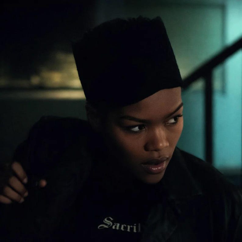 May include: A person wearing a black hat and a black shirt with the word "Sacril" in white script. The person is looking off to the side with a serious expression. The background is dark and out of focus.