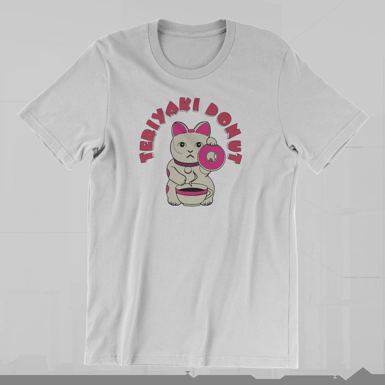 May include: White t-shirt featuring a cartoon cat holding a pink donut and a cup. The text "TERIYAKI DONUT" arches above the cat in pink letters. The cat has pink ears and spots, and a serious expression.