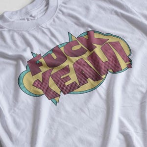 Puede incluir: Camiseta blanca con un estampado gráfico de las palabras "F**K YEAH!" en un estilo de cómic con colores amarillo, rosa y azul.