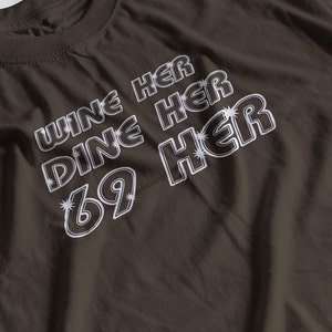 May include: A brown t-shirt with white text that reads "Wine Her Dine Her 69 Her".