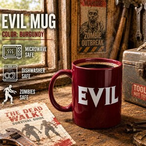 Op de afbeelding: Een bordeauxrode keramische mok met het woord EVIL in grote witte hoofdletters op de voorzijde. De mok is gevuld met zwarte koffie en bevat iconen die aangeven dat deze magnetronbestendig, vaatwasserbestendig en zombieveilig is.