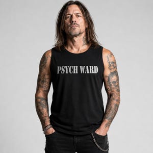 Op de afbeelding: Zwart mouwloos shirt met de tekst "PSYCH WARD" in het wit. De persoon die het shirt draagt heeft tatoeages op zijn armen en nek. Ze dragen een zwarte jeans. De achtergrond is effen grijs.