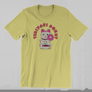 May include: A light yellow t-shirt featuring a cartoon cat holding a pink donut and a cup. The text "TERIYAKI DONUT" arches above the cat in pink letters. The cat has gray fur with pink accents.