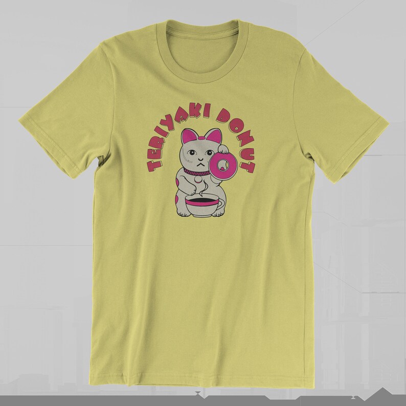 May include: A light yellow t-shirt featuring a cartoon cat holding a pink donut and a cup. The text "TERIYAKI DONUT" arches above the cat in pink letters. The cat has gray fur with pink accents.