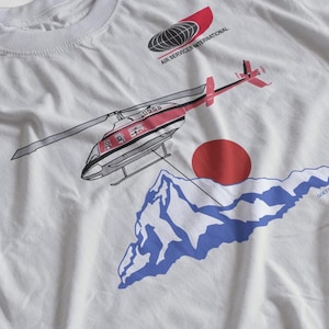 May include: A white t-shirt with a red and black helicopter graphic, a red sun, and blue mountains. The text "AIR SERVICES INTERNATIONAL" is printed on the shirt.