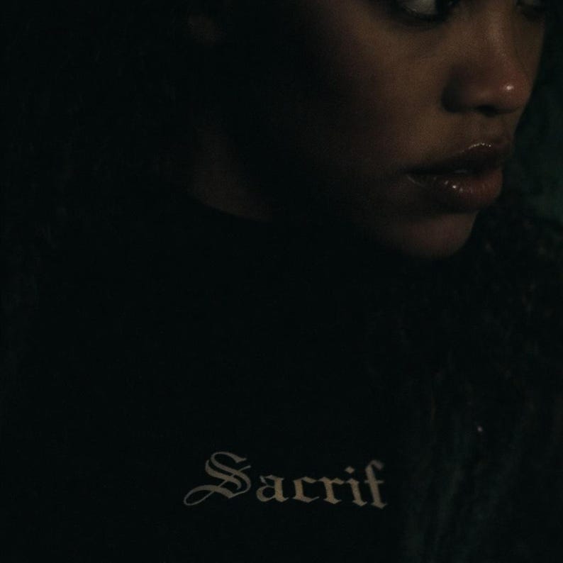 May include: A black turtleneck top with the word "Sacrif" in a gothic font. The text is in a lighter colour, contrasting against the dark fabric. The image is a close-up, focusing on the shirt and the text.