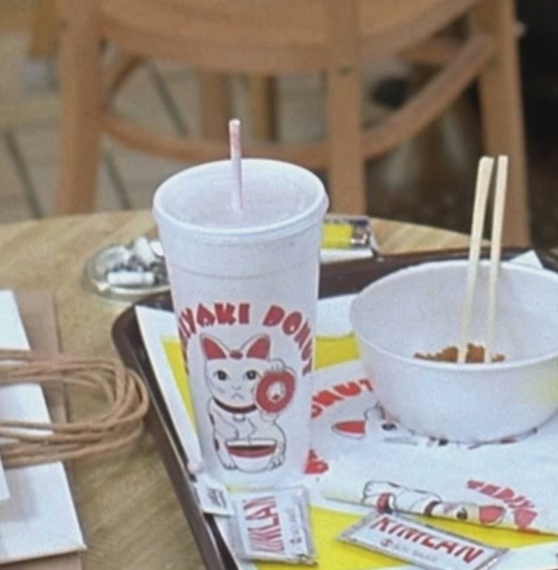 May include: A white paper cup with a pink straw, featuring the text "Yaki Donut" and a lucky cat illustration. The cup sits on a tray with a bowl of food and chopsticks. Small packets of sauce are also visible.