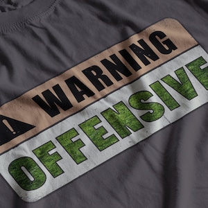 Puede incluir: Una camiseta gris oscuro con un cartel de advertencia que dice "WARNING OFFENSIVE" en letras negras y verdes. El cartel tiene un fondo amarillo con un signo de exclamación negro.