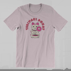 May include: A light pink t-shirt featuring a cartoon cat holding a pink donut and a cup of coffee. The text "TERIYAKI DONUT" arches above the cat in pink letters. The cat has pink ears and spots.
