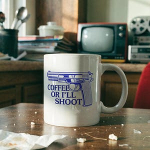 以下が含まれることがあります： 青い拳銃のグラフィックと青い文字でCOFFEE OR I'LL SHOOTとプリントされた白いセラミック製のマグカップ。マグカップは標準的な円筒形で、右側に湾曲したハンドルが付いており、鮮明で大胆なデザインが特徴です。