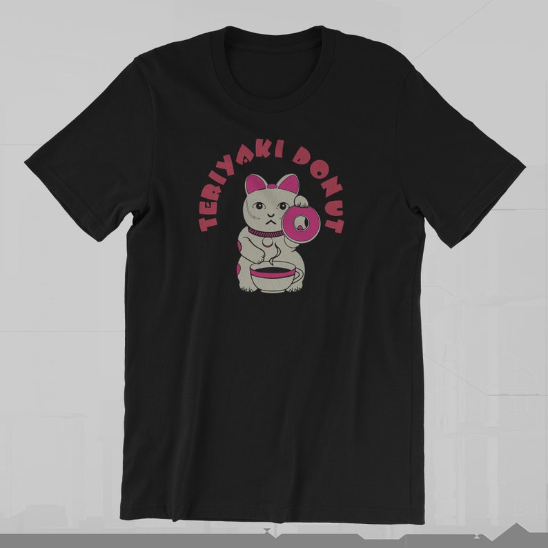 May include: Black t-shirt featuring a cartoon cat holding a donut and a cup of coffee. The text "TERIYAKI DONUT" is printed above the cat in a pink, circular design. The cat has gray fur and pink accents.