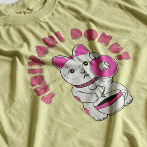 May include: A light yellow t-shirt featuring a cartoon cat holding a pink donut and a cup of coffee. The text "TERIYAKI DONUT" arches above the cat in pink letters. The cat has pink accents on its ears and donut.