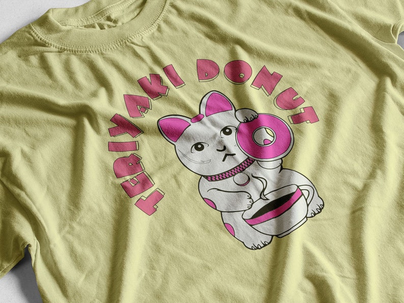 May include: A light yellow t-shirt featuring a cartoon cat holding a pink donut and a cup of coffee. The text "TERIYAKI DONUT" arches above the cat in pink letters. The cat has pink accents on its ears and donut.
