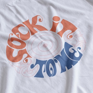 以下が含まれることがあります： 白いTシャツに、レトロなサイケデリックなフォントで「Sock It To Me」という文字が円形にデザインされています。文字はオレンジとブルーの色合いです。