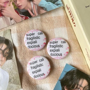 Puede incluir: Tres chapas circulares con fondo blanco y un patrón festoneado rosa claro. Cada chapa muestra el texto negro supercalifragilisticexpialidocious dispuesto en cinco líneas. Las chapas están colocadas en una formación triangular.