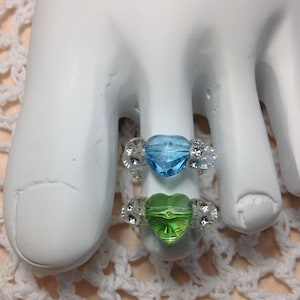Puede incluir: Dos anillos para los dedos del pie con cristales en forma de corazón. El anillo superior tiene un cristal azul claro y el anillo inferior tiene un cristal verde claro. Ambos anillos están decorados con diamantes de imitación transparentes.