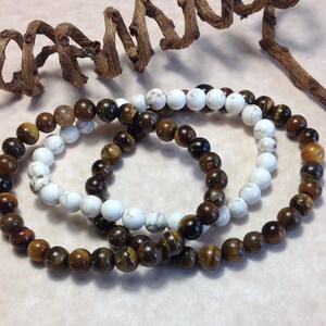 May include: Three bracelets made of brown tiger's eye beads and white beads. The bracelets are stacked on top of each other.
