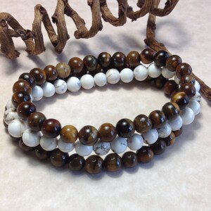 May include: Two beaded bracelets, one brown and gold tiger's eye and one white, are stacked on top of each other.