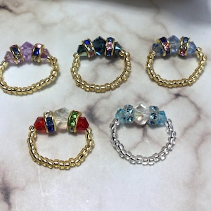 Puede incluir: Cinco anillos de cuentas con cristales de diferentes colores. Los anillos están hechos con cuentas doradas y plateadas. Los cristales son en tonos de azul, rosa, rojo y verde.