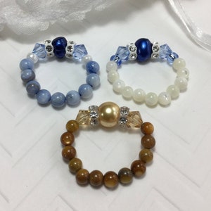 May include: Three beaded rings with different colored stones. The first ring has blue and white beads with a blue pearl in the center. The second ring has white beads with a blue pearl in the center. The third ring has brown beads with a gold pearl in the center.