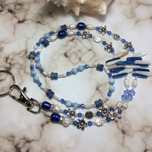 Puede incluir: Un cordón con cuentas con un cierre plateado, con cuentas azules, blancas y color perla. El cordón incluye dijes de flores plateadas y un grupo de acentos azules y blancos. Una concha marina descansa en el fondo.