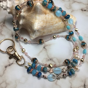 May include: A multi-strand beaded lanyard with a gold key ring. The lanyard features blue, turquoise, and gold beads, as well as small pink and white beads. The lanyard is draped over a seashell.