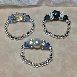 Puede incluir: Tres anillos de cuentas plateadas con diferentes centros. El primer anillo tiene un cristal transparente iridiscente, el segundo anillo tiene una cuenta negra y el tercer anillo tiene una perla blanca.