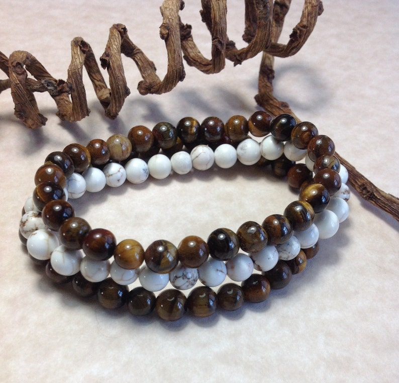 May include: Two beaded bracelets, one brown and gold tiger's eye and one white, are stacked on top of each other.