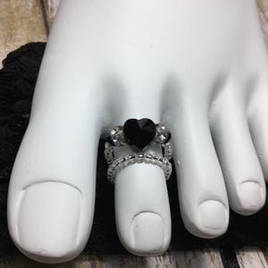Puede incluir: Un anillo de dedo del pie de cristal negro en forma de corazón con detalles de cristal transparente. El anillo está hecho de metal plateado y se lleva en el dedo del pie de una modelo.