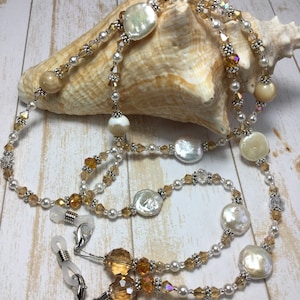 May include: A double-strand glasses chain with gold and white beads. The chain features large, round, white mother-of-pearl beads and smaller, faceted, gold-toned beads. The chain is attached to clear plastic eyeglass holders.