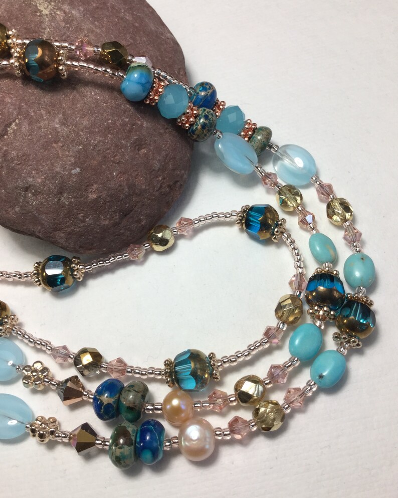 May include: A multi-strand necklace with turquoise, blue, and gold beads. The necklace is made with a variety of materials, including glass, metal, and stone.