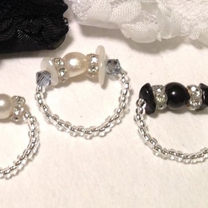 Puede incluir: Tres anillos de plata con diferentes diseños de cuentas. El primer anillo tiene dos cuentas blancas y una de color melocotón. El segundo anillo tiene una perla blanca, un botón blanco y un cristal transparente. El tercer anillo tiene una cuenta negra y dos cristales transparentes.