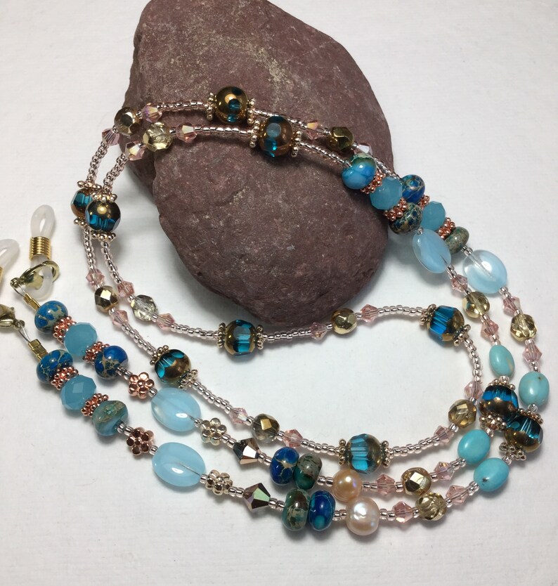 May include: A multi-strand beaded eyeglass chain with blue, turquoise, and gold beads. The chain is made of small beads and larger, oval-shaped beads. The chain is attached to two small gold clips.