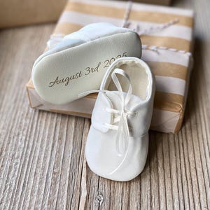 Puede incluir: Un par de zapatos de bebé blancos con cordones de cinta de satén. Un zapato está colocado en posición vertical, mientras que el otro está de lado, revelando el texto "August 3rd 2025" impreso en una fuente cursiva oscura en la suela.