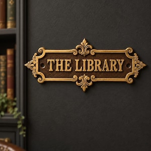 May include: A rectangular wall plaque with a dark brown textured background and raised gold-toned lettering that reads THE LIBRARY. The plaque features ornate scrollwork and fleur-de-lis accents on the edges, with two mounting holes on the sides.