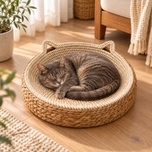 Pode incluir: Uma cama redonda para gatos, tecida em fibra natural de cor castanha clara, com uma borda elevada que apresenta dois pequenos detalhes em forma de orelhas pontiagudas. Um gato cinzento tigrado dorme enrolado no centro sobre tecido claro.