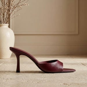 May include: A deep burgundy leather mule sandal featuring a pointed toe and a slender, flared stiletto heel. The shoe has a single wide strap across the front, providing an open-back design with a smooth, solid-colored finish throughout the exterior.