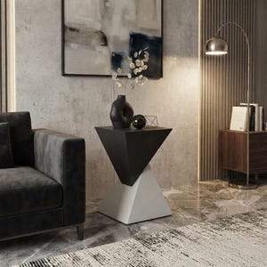 Peut inclure: Une table d'appoint géométrique composée d'une section supérieure triangulaire noire et d'une base triangulaire blanche. La table est placée à côté d'un canapé gris foncé et porte un vase noir avec des tiges séchées et une petite sphère noire.