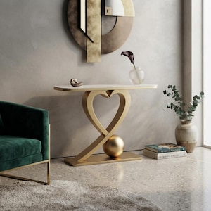 May include: A console table featuring a white rectangular top supported by a gold-colored base shaped like a heart with a solid gold sphere nestled at the bottom. The table stands on a flat rectangular base, providing a stable foundation for the structure.
