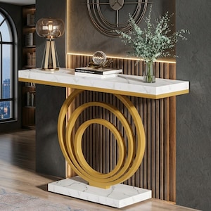 May include: A console table featuring a white faux marble rectangular top and base with gold trim. The support structure consists of three concentric gold metal circles. A glass lamp, a stack of books, and a glass vase with greenery sit on top.