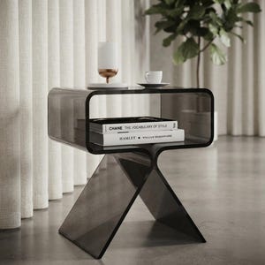 Peut inclure: Une table d'appoint en acrylique gris foncé translucide avec un cadre incurvé et continu. La table dispose d'une étagère centrale contenant deux livres intitulés CHANE THE VOCABULARY OF STYLE et HAMLET WILLIAM SHAKESPEARE, surmontés d'une bougie et d'une tasse blanches.