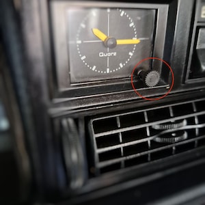 May include: A black analog quartz dashboard clock with yellow hour and minute hands, white tick marks, and the word "Quarz" printed on the face. A small, circular, textured black adjustment knob is positioned on the lower right side of the clock.