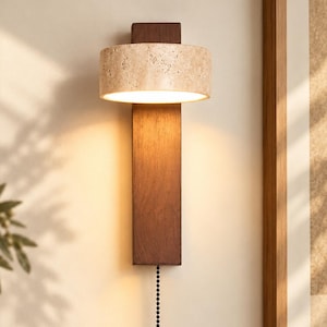 May include: A wall-mounted light fitting featuring a vertical dark brown wooden rectangular backplate and a cylindrical beige stone shade. The lamp emits a warm glow and includes a black beaded pull chain hanging from the bottom of the wood.