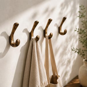 May include: Four wall-mounted hooks with a brushed antique brass finish. The hooks feature a curved, J-shaped design. A beige textured towel hangs from the second hook, and a beige canvas tote bag hangs from the third hook on the wall.